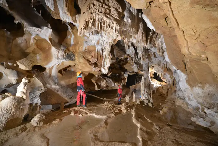 Exploratrice vêtue de rouge et portant un casque avec une lampe frontale, accroupie dans une grotte devant une formation rocheuse d'où jaillit de l'eau scintillante.